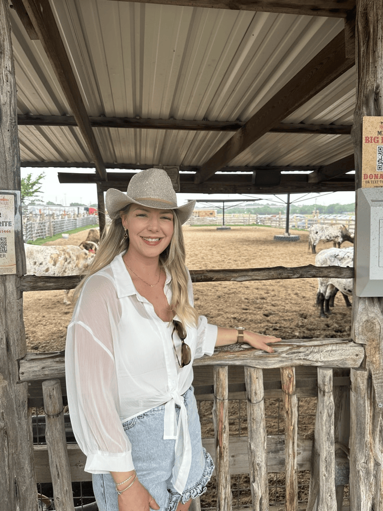 A woman wearing a white shirt, denim shorts, and a beige hat stands by a wooden fence at a farm, smiling at the camera with animals in the background.