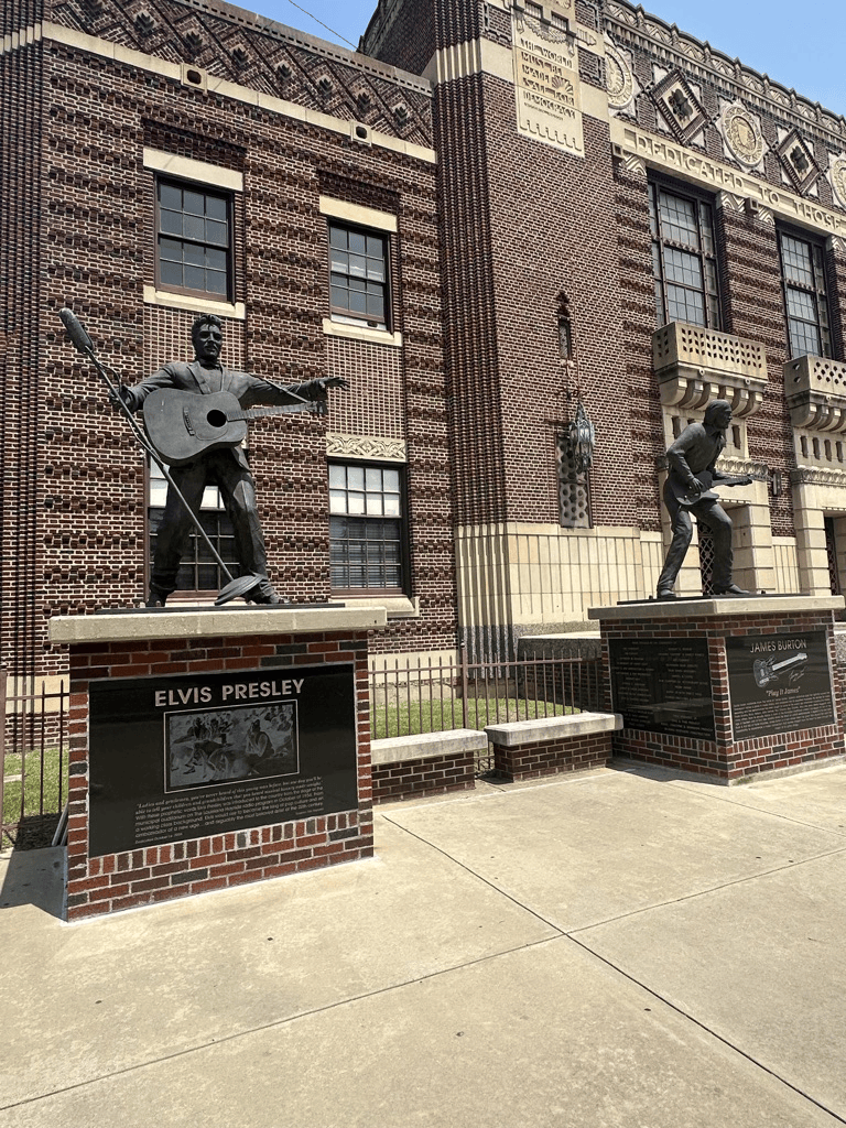 Bronze statues of Elvis Presley and James Burton outside a brick building, with information plaques at their bases.
