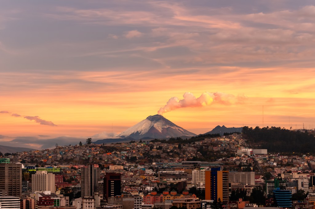 View of a city skyline at sunset with a snowy volcano emitting smoke in the background.