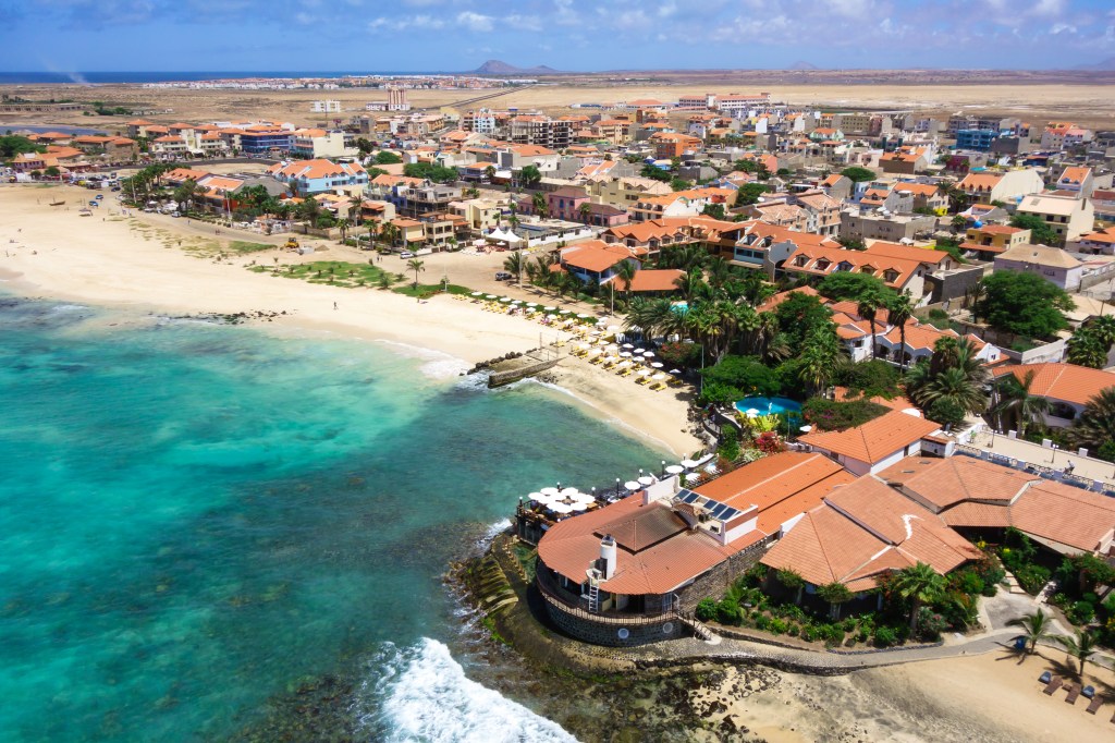 Aerial view of a coastal town featuring sandy beaches and colorful buildings. The turquoise water meets the shore, with palm trees and lounge chairs visible along the beach.