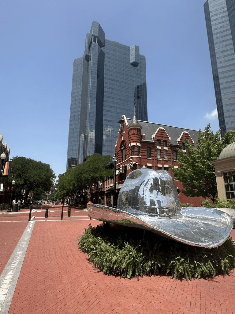 Giant metallic cowboy hat sculpture surrounded by greenery and located on a red brick street, with modern skyscrapers in the background.