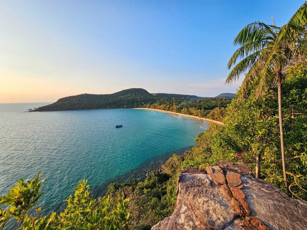 A scenic view of a calm sea and sandy beach surrounded by lush greenery and hills under a clear blue sky.